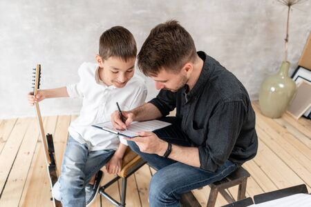 Adult man explains his son, who learn to play guitar, how to play melodies and songs using music sheet. Education with familyの写真素材