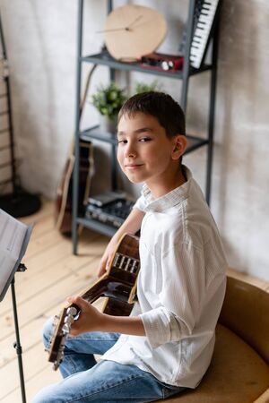 Young boy in casual clothing sits on chair with acoustic guitar in hands and posingの写真素材