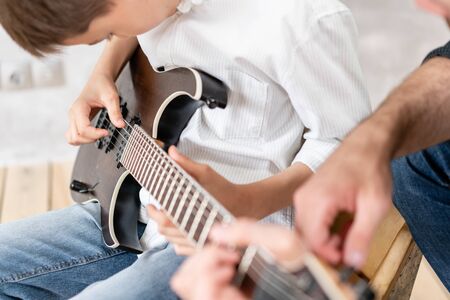 Young boy moves his hand along guitar strings while his father helps boy to take chordsの写真素材