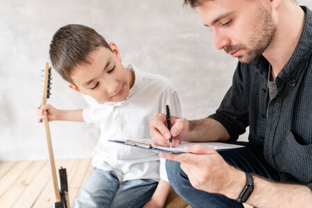 Adoult man writes notes of simple melody on music sheet for his younger brother, who learn to play guitarの写真素材