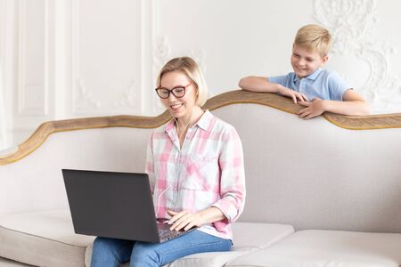 Beautiful woman sitting on couch and her son standing behind it. They talking with relatives through laptop.の写真素材
