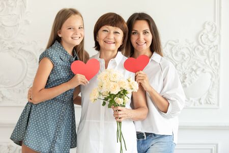 Mother, daughter and granddaughter hug and hold cardboard hearts and a bouquet of flowers