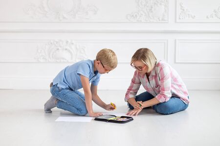 A young tutor in a plaid shirt and her young student, wearing a blue t-shirt and glasses, sit on a white floor. The woman watches as the boy drawsの写真素材
