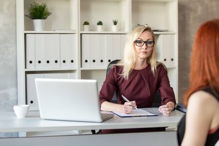 Blond woman in glasses sits behind office table and listen other woman. Selective focusの写真素材