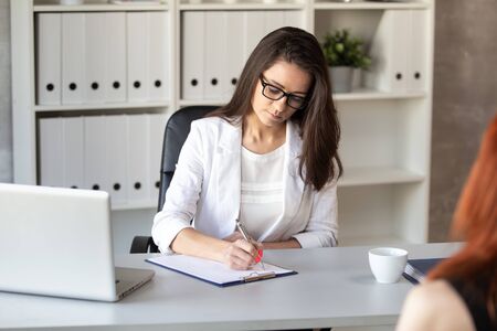 Woman in white jacket sits behind office table and doing notes in sheet of paper. Selective focusの写真素材