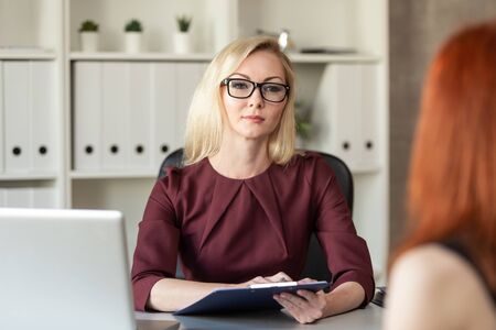 Blond woman in glasses sits behind office table and listen other woman. Selective focusの写真素材