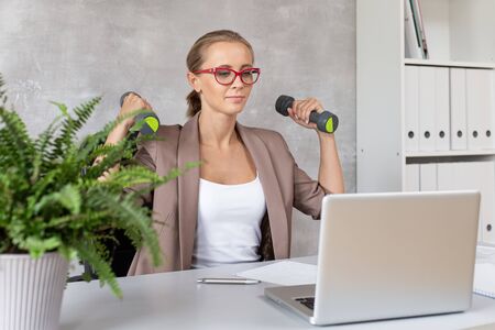 Smiling woman sits in chair with two dumbbells in hands and looking at laptopの写真素材