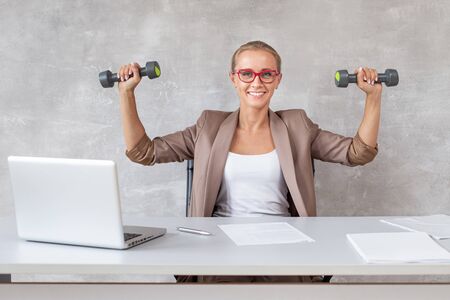 Blond office worker sits in chair behind working desk and holding two dumbbells in handsの写真素材