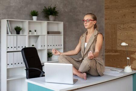 Cute businesswoman in yoga pose relaxing on table in officeの写真素材