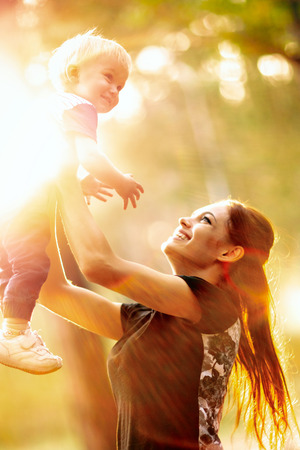 Young mother holds her son. Woman is having fun with her little baby in a summer park. Happy mom throws up child while playing outdoorsの写真素材
