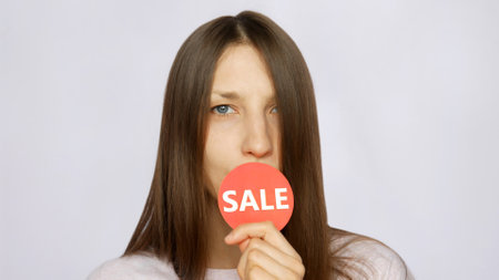 A young woman holding a red round sign with the word SALE in her hand and leaning it to her lipsの写真素材