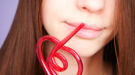 One beautiful girl drinks from a pink heart-shaped straw close-upの写真素材