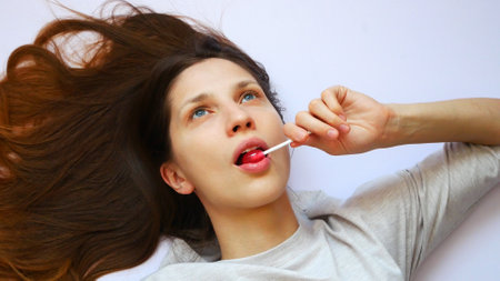 A very beautiful long-haired girl lies on a white surface and sucks a red lollipop dreamingの写真素材