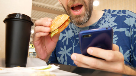 Close-up of a man using a smartphone and eating a hamburger in a dinnerの写真素材