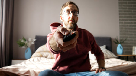 A man with a remote control in his hand changes TV channels sitting on a bedroom bed