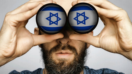 The Israeli flag is reflected in the lenses of binoculars through which a bearded man looks