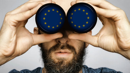 The European union flag is reflected in the lenses of binoculars through which a bearded man looks