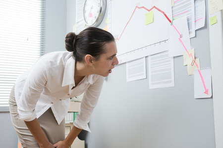 Stunned businesswoman checking a financial business chart on office wall with arrow going down.の写真素材