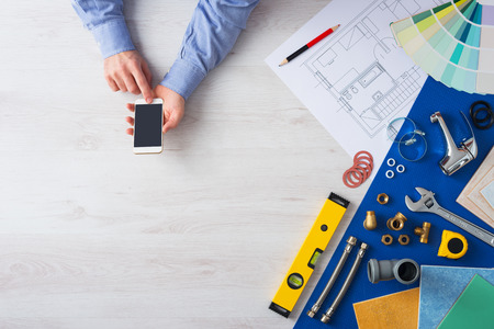 Male hands using a mobile phone next to plumbing work tools, tiles and swatches, online booking and...