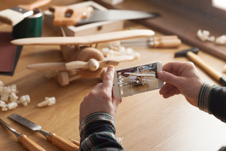 Man photographing his handmade wooden toy airplane with a smart phone on a work tableの写真素材