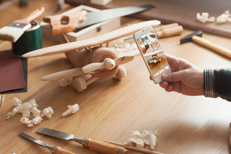 Man photographing his handmade wooden toy airplane with a smart phone on a work tableの写真素材