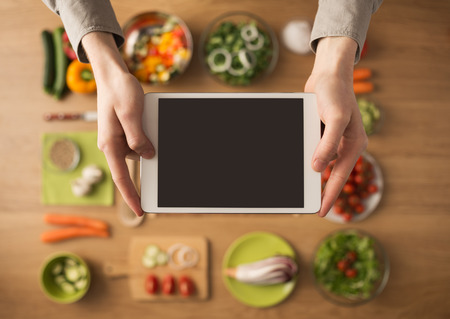 Hands holding a digital touch screen tablet with fresh vegetables and kitchen utensilsの写真素材