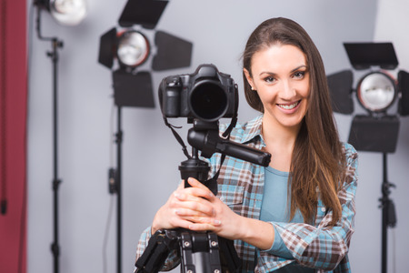 Young female photographer posing in her studio with a digital camera on a tripod and lighting...