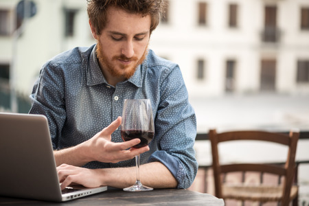 Young  man sitting at outdoor bar table and smelling a glass of red wineの写真素材