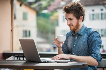 Young man connecting with a laptop at the bar and drinking a cappuccino, town buildings on backgroundの写真素材
