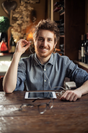 Cool hipster sitting at bar table and listening to music with his tablet using earphonesの写真素材