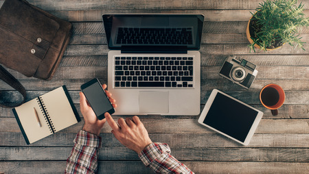 Vintage hipster wooden desktop top view with laptop, vintage camera, plant and touch screen tablet, male hands using a mobile phoneの写真素材