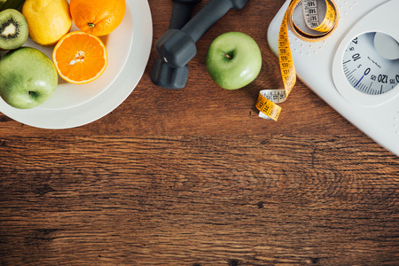 Fitness and weight loss concept, dumbbells, white scale, fruit and tape measure on a wooden table, top view