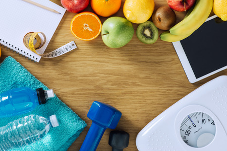 Fitness and weight loss concept, dumbbells, white scale, towels, fruit, tape measure and digital tablet on a wooden table, top view