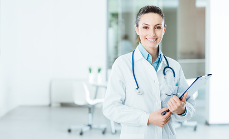 Smiling female doctor with lab coat in her office holding a clipboard with medical records, she is looking at camera Smiling female doctor with lab coat in her office holding a clipboard with medical records, she is looking at camera