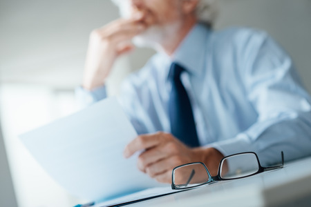 Pensive businessman with hand on chin looking away and holding a document, selective focus, glasses on foreground