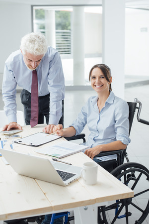 Confident business woman in wheelchair working at office desk with her male colleague and smiling at cameraの写真素材