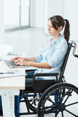 Young disabled business woman in wheelchair working at office desk and typing on a laptop, accessibility and independence conceptの写真素材