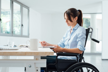 Confident disabled business woman in wheelchair working at office desk and checking paperworkの写真素材
