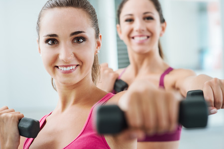 Cheerful young women exercising at the gym and weightlifting using dumbbells, they are smiling at cameraの写真素材