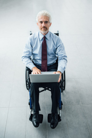 Confident smiling businessman in wheelchair using a laptop, office interior on backgroundの写真素材