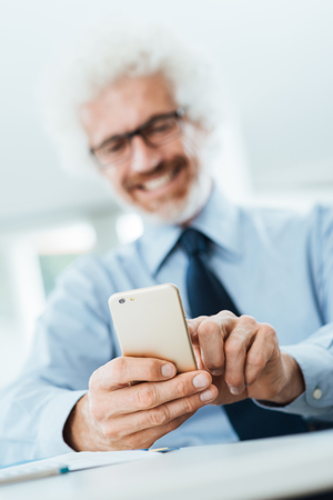 Smiling businessman sitting at office desk and using a touch screen smart phone, texting and networking onlineの写真素材