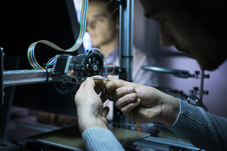 Young engineer in the lab working on a 3D printer, another student is working with a computer on...