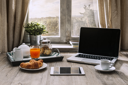Laptop and coffee on a wooden table in front of a window in a rustic house, hipster breakfast conceptの写真素材