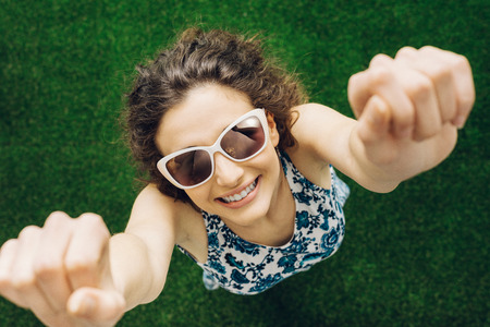 Cheerful smiling young woman standing with raised fists and stretching on the grass, she is wearing sunglassesの写真素材