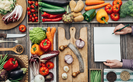 Freshly harvested vegetables, cooking utensils and chopping boards on a rustic kitchen worktop, a cook is writing recipes on a notebookの写真素材