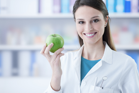 Female dentist smiling and holding a green apple, dental care and prevention conceptの写真素材