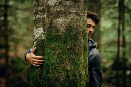 Young man relaxing in the forest and hugging a moss covered treeの写真素材