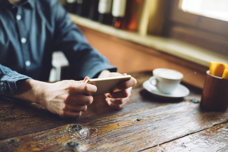 Young hipster guy texting with his mobile phone at the bar and having a cappuccinoの写真素材