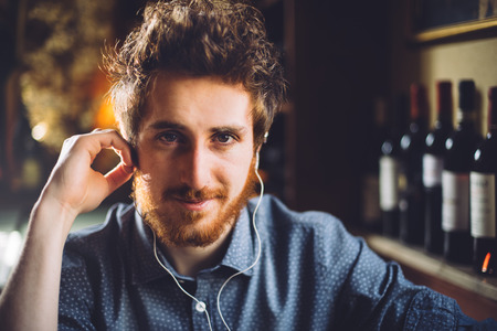 Young hipster man sitting at bar table and listening to music using earphones, he is smiling at...