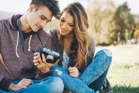 Young couple sitting on the grass in the sunlight and watching pictures on a digital camera,...